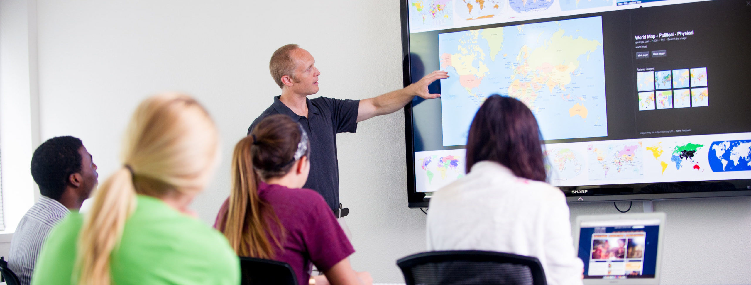 Man teaching students pointing to projector screen displaying world maps.