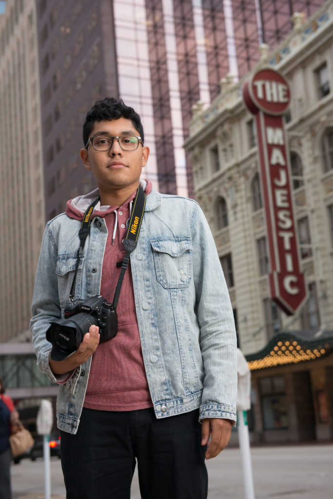 Young man with camera in hand surrounded by tall buildings and a Majestic Theater sign in the background.