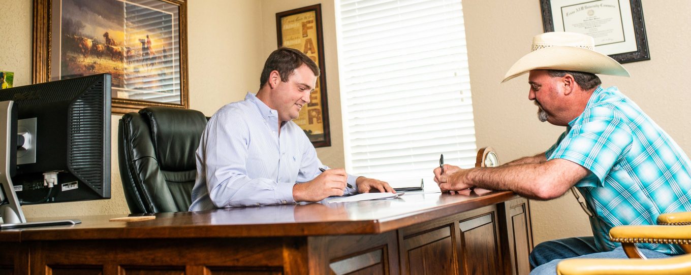 Two men signing documents in an office, one is sitting behind the desk with a computer on his side and the other is sitting in front wearing a hat. 