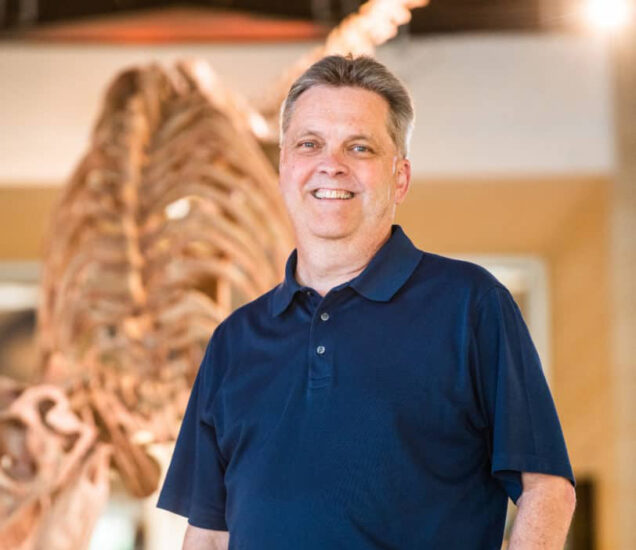 Dr.-Donham protrait in front of a dinosaur structure.