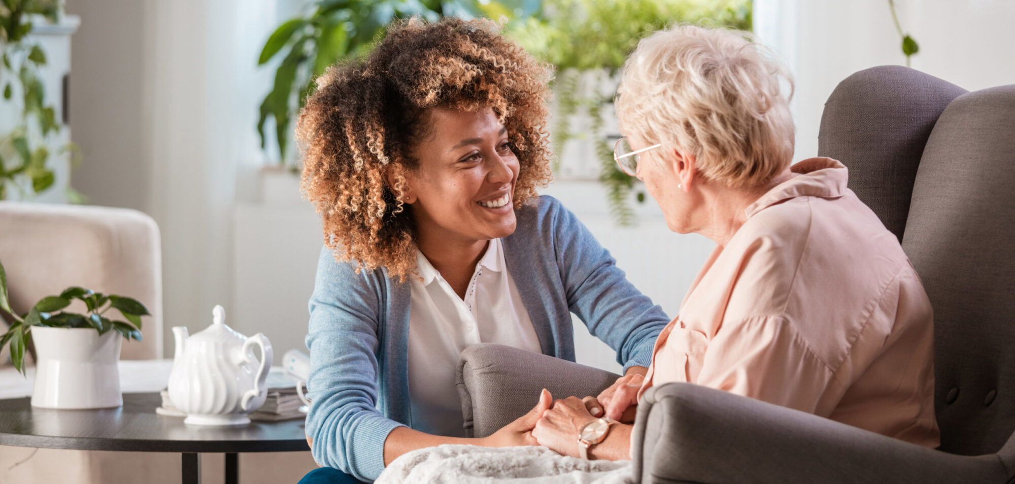 Female home caregiver talking with senior woman, sitting in living room and listening to her carefully.