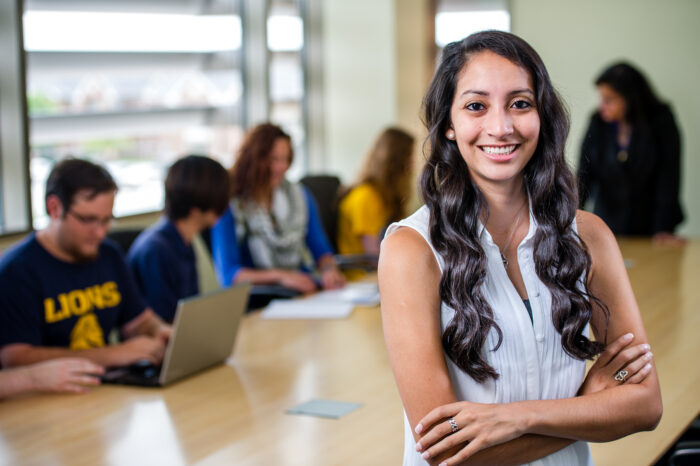 Student smiling posing for photo in front of a desk where other students are sitting down working together.