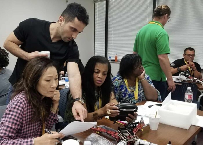 A group of people are gathered around a table, focused on a project involving electronics. One person is pointing at a document while others examine various components. The room appears to be a workshop or training session, with tools and materials scattered on the table.