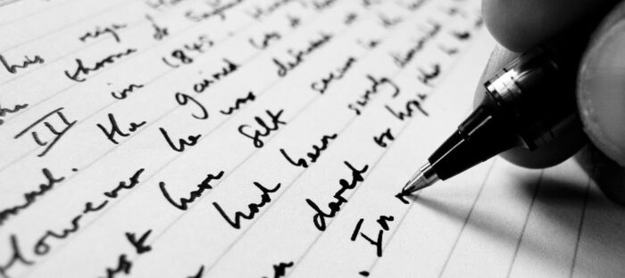 Person writing on a sheet of paper with a pen, zoomed in the fingers holding the pen and the words on the paper.