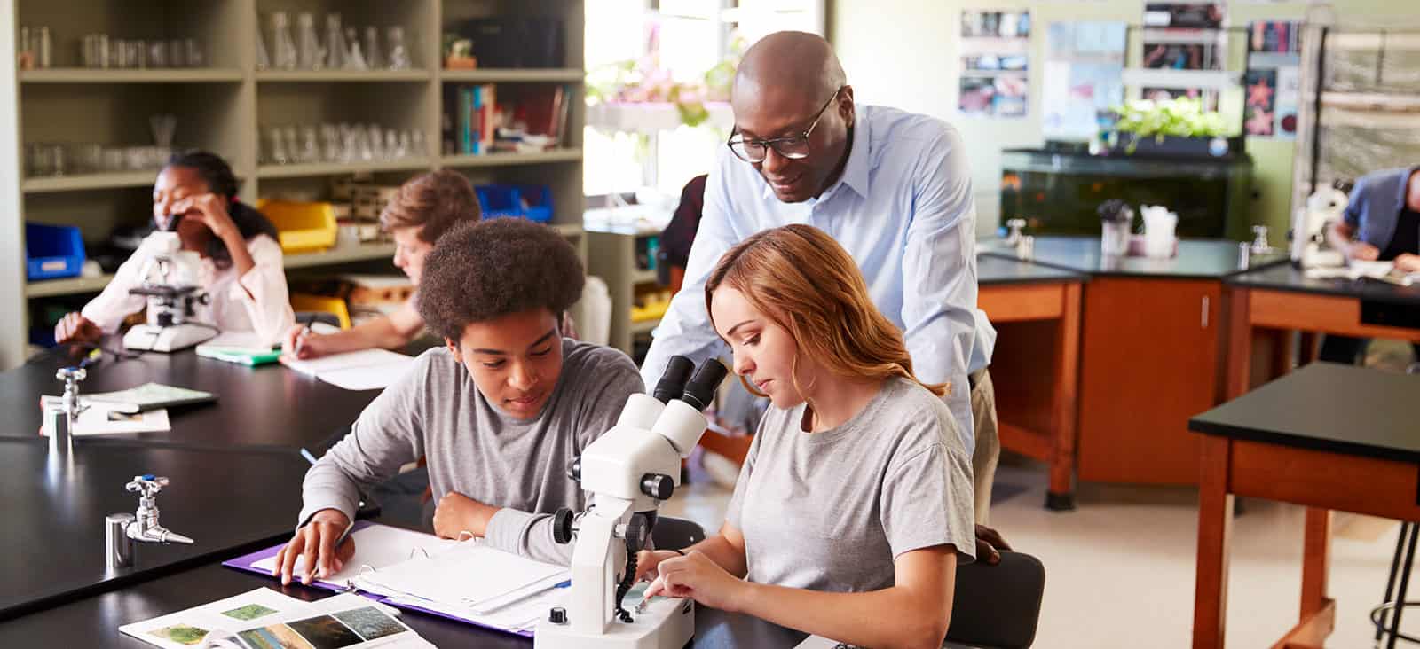 High School Students With Tutor Using Microscope In Biology Class