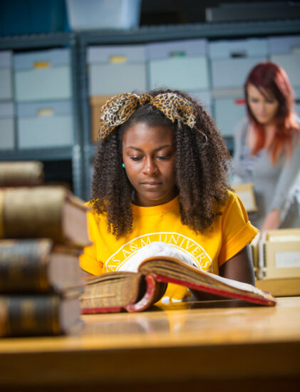 Female student looking at old hardback book with another female student in the background