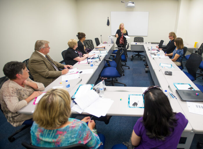 A group of professors sitting down at a table listening to one professor standing in front of them.