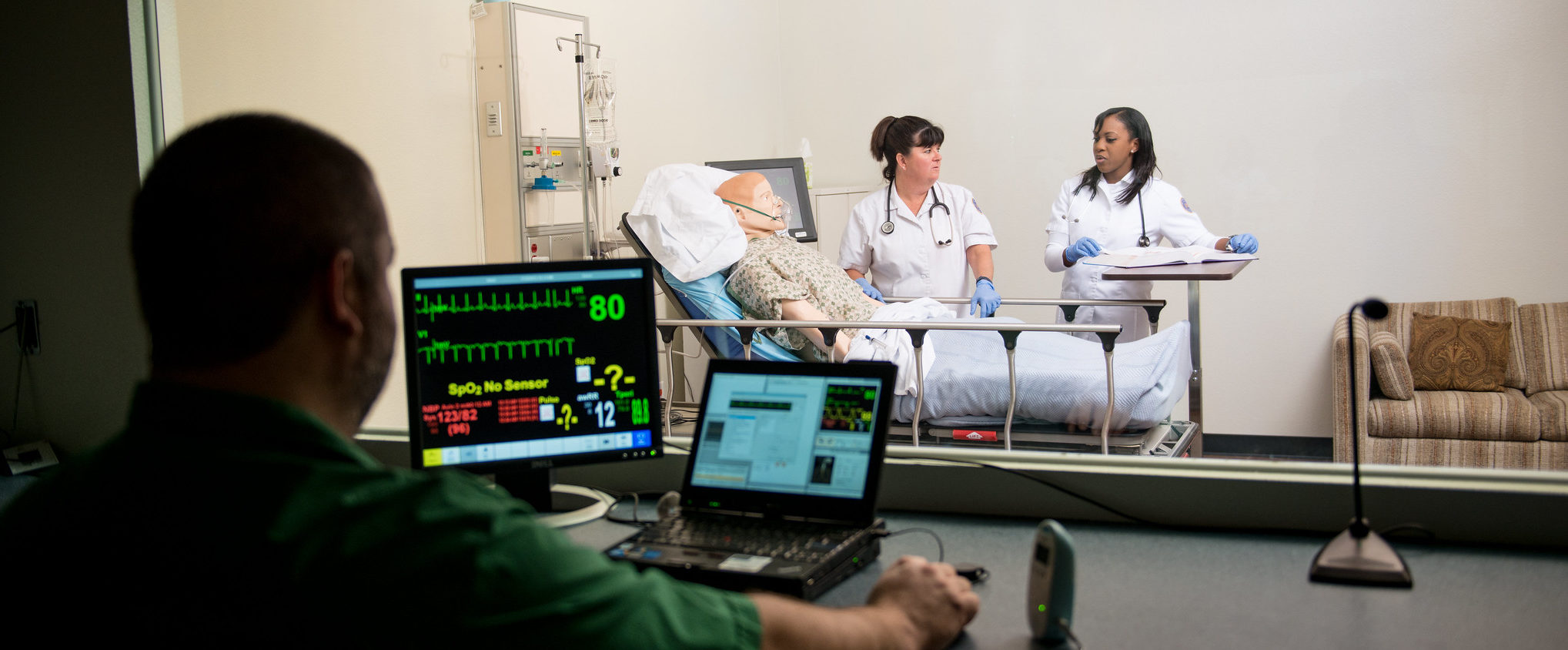 A man at computer behind computers with viewing window of nurses practicing on a mannequin.