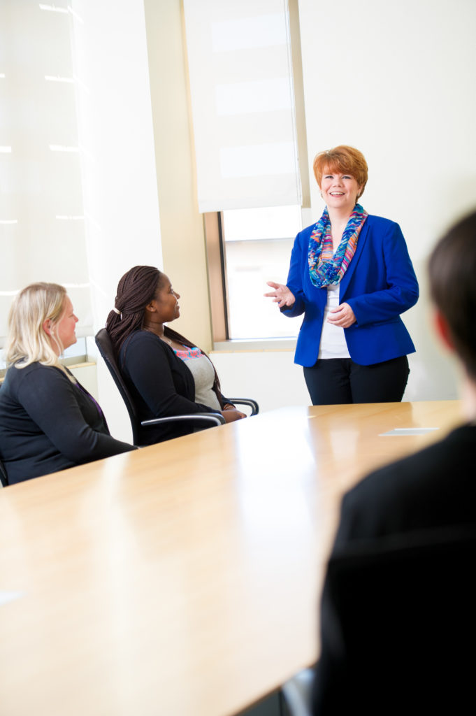 Business dressed woman speaking to group around a large table.