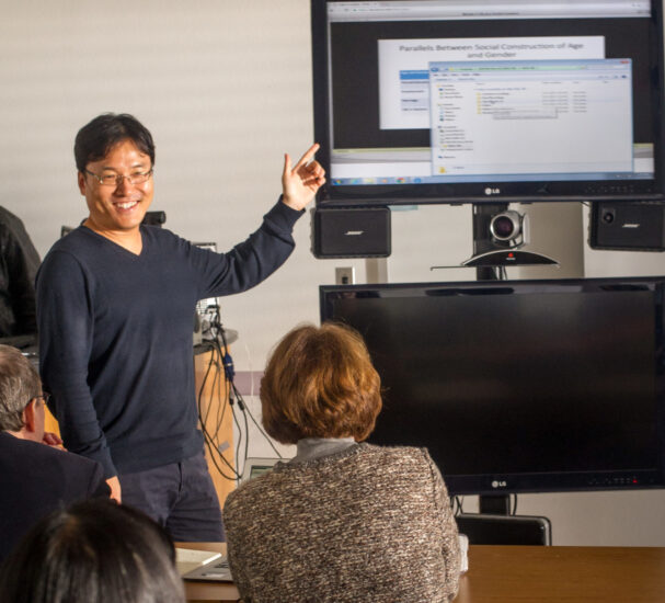 Male teacher point at TV screen and conferencing camera in front of class.