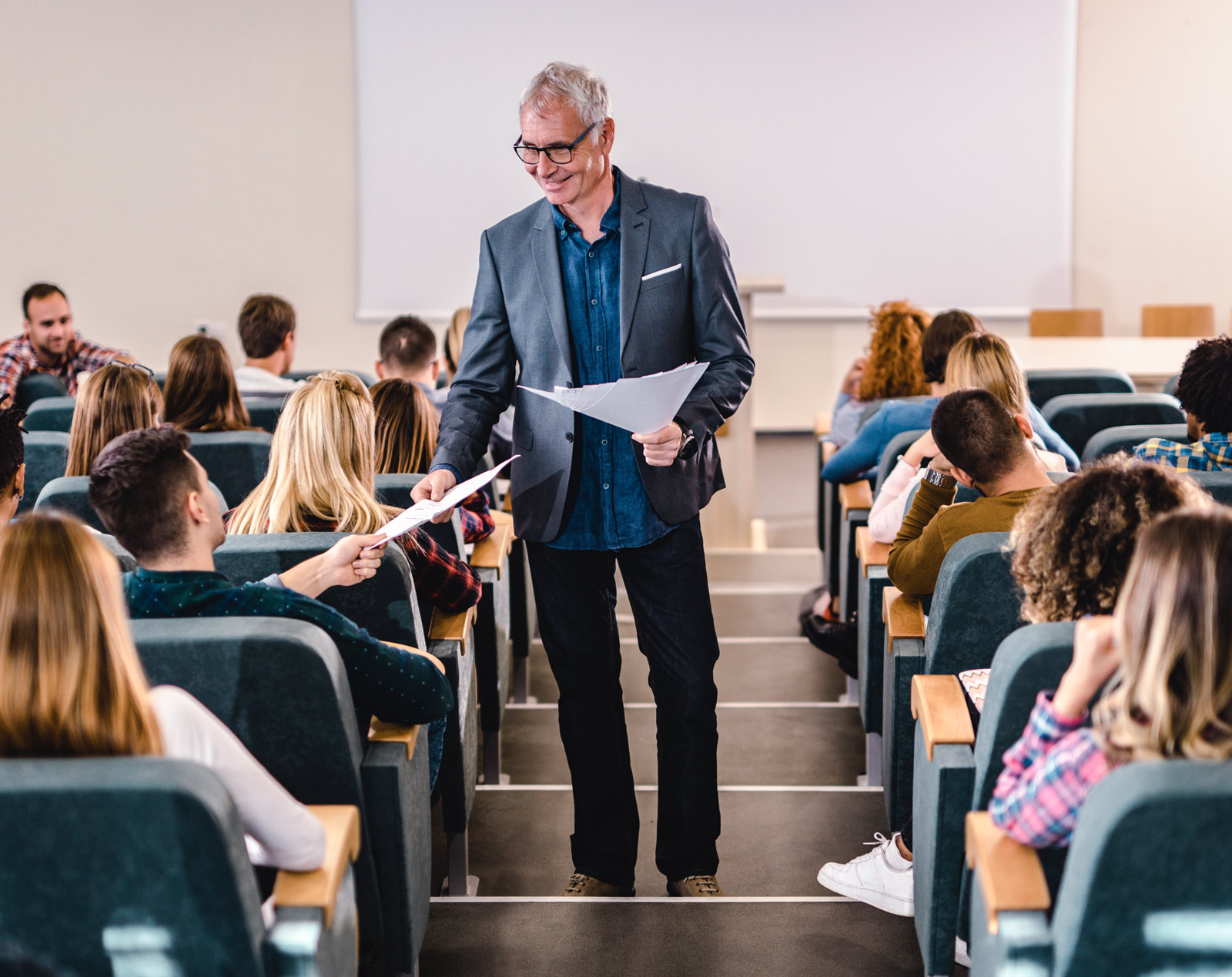 Happy senior professor talking to his students while giving them test results in lecture hall.