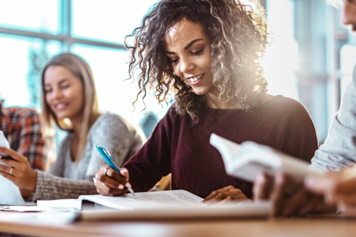 Female student at desk reading a book with pen in hand.