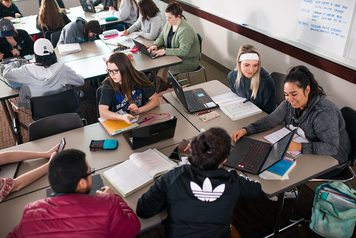 A group of students sitting down in class facing each other, using computers and talking.