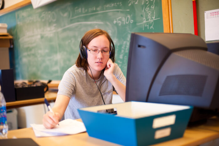 A young woman writing on paper while listing to headphones at a computer.