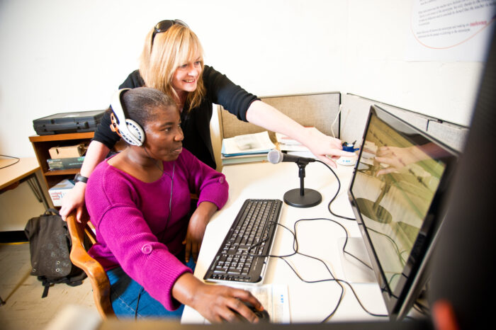 An educator teaching a student who is setting at a computer while wearing headphones.