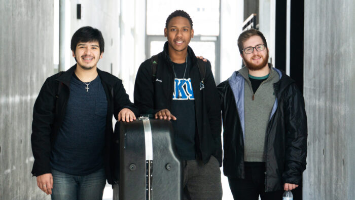 Three young men standing in hallway with large instrument in case