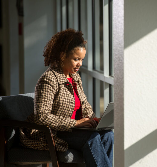 Woman working on laptop seated facing windows.