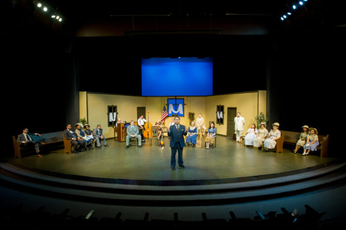 Lead actor standing on stage front with actors behind.