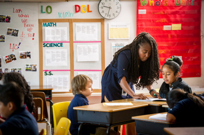 Woman teacher readying notes book on desk surrounding by young students.