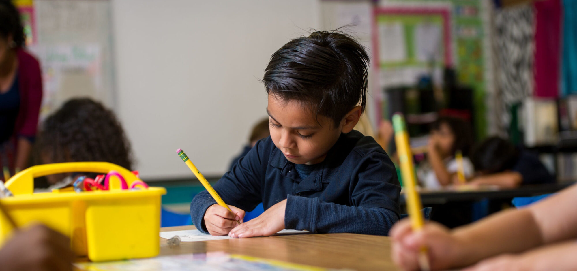 Young hispanic boy writing with pencil in classroom.