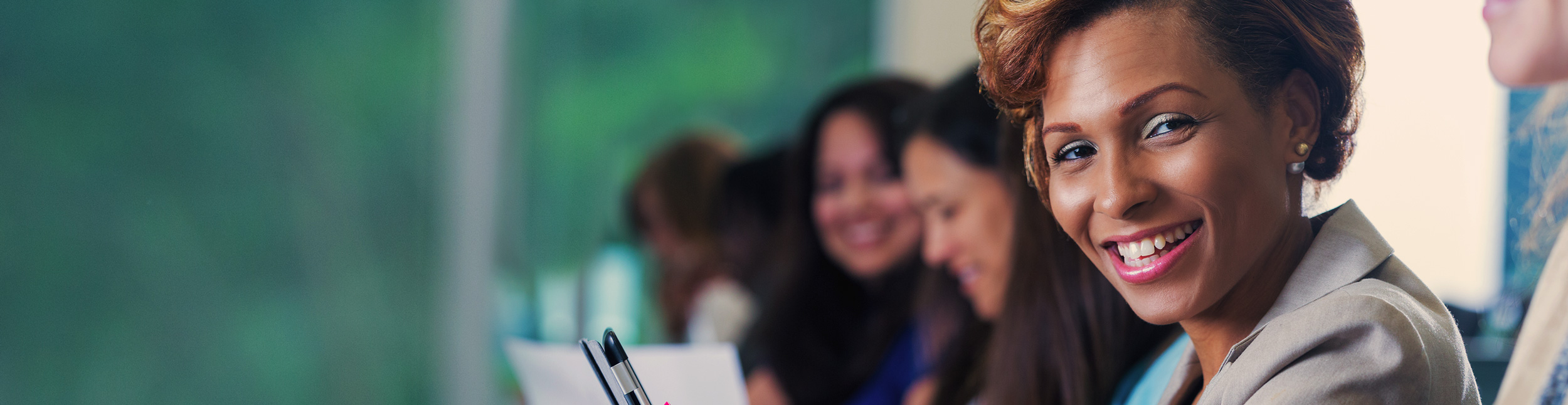 Mid adult African American businesswoman is smiling and looking at the camera. She is using her smart phone to take notes while listening to a speaker during a business conference or job training seminar. She's sitting in a lecture hall with diverse professional colleagues.