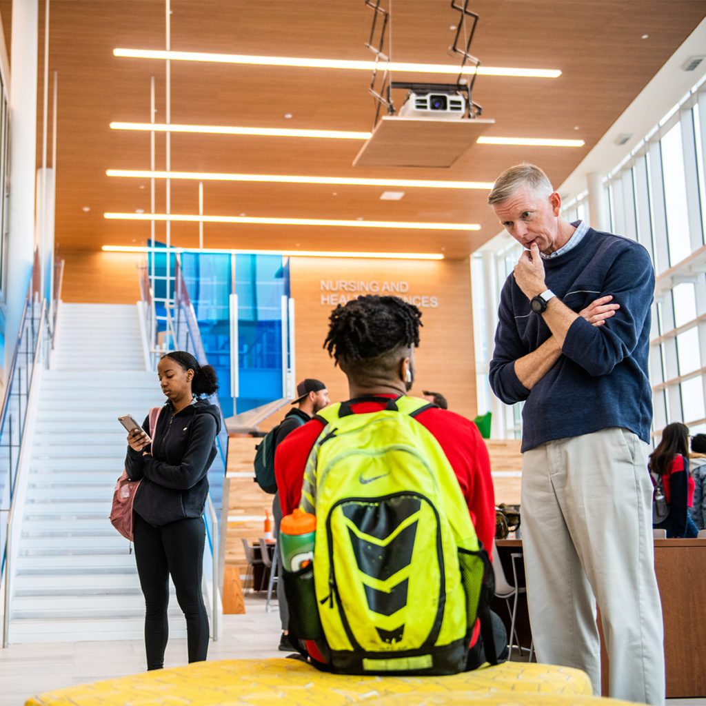 A professor talking to a student in the common area of the nursing and health sciences building.
