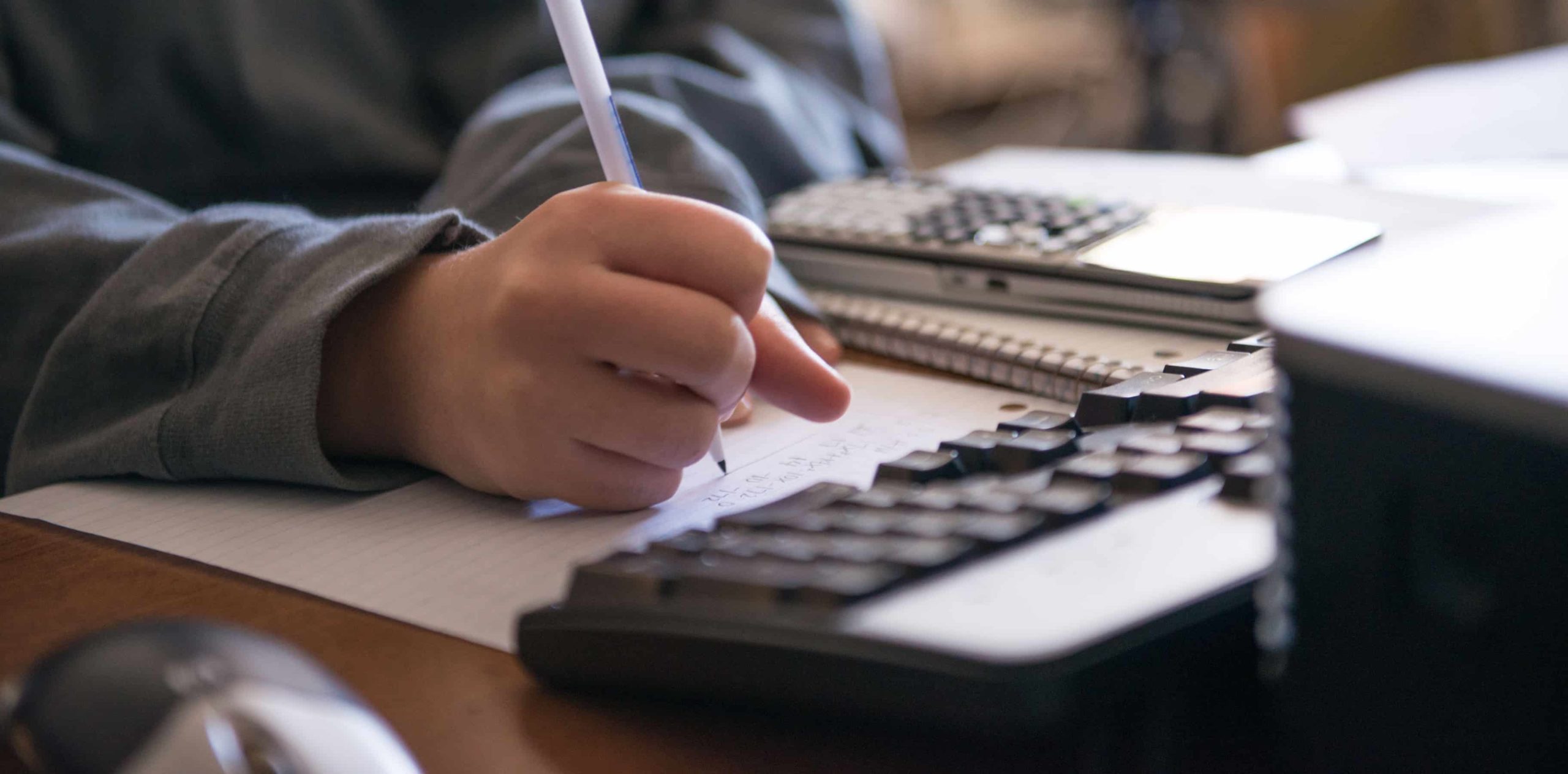 Someone appearing to do a math problem on a sheet of paper with a computer in front of them and a calculator on the side.