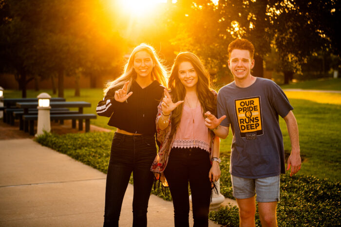 three students making the TAMUC sign while taking a picture on campus with the sunset behind them.