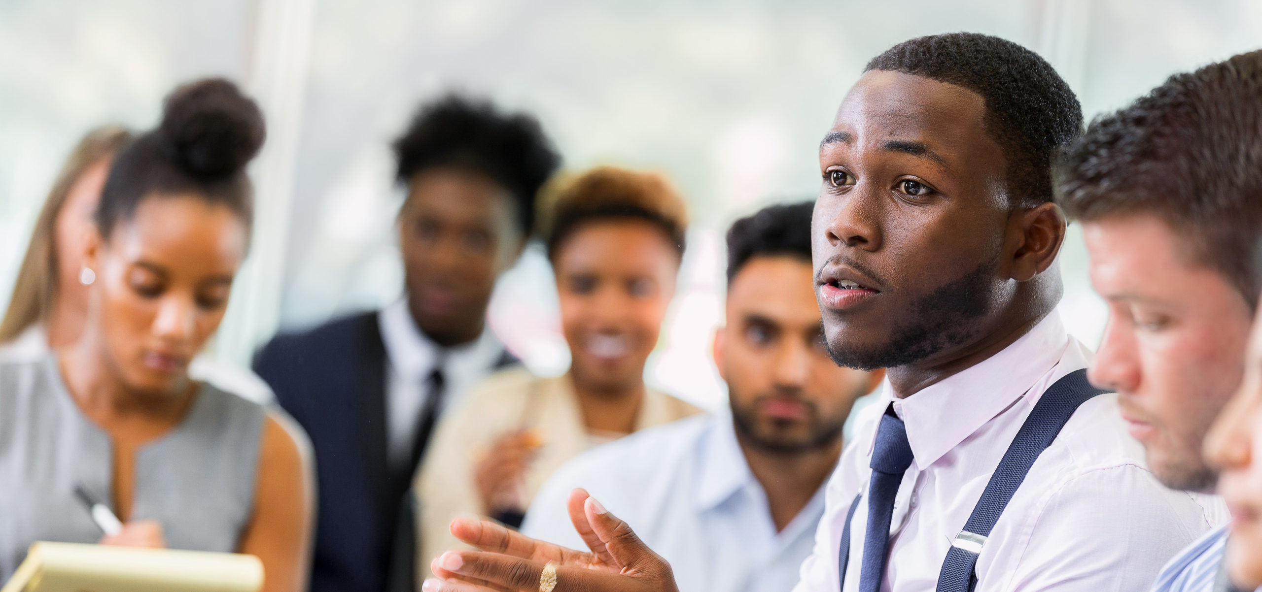 A serious young African American male business student talks with his hands as he sits at a table in a lecture hall and poses a question to an unseen professor. Other students sit in the audience in the background.