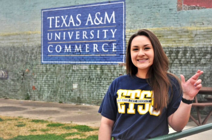 Picture of Heather Rodriguez making the lion sign with behind a mural of Texas A&M University Commerce.