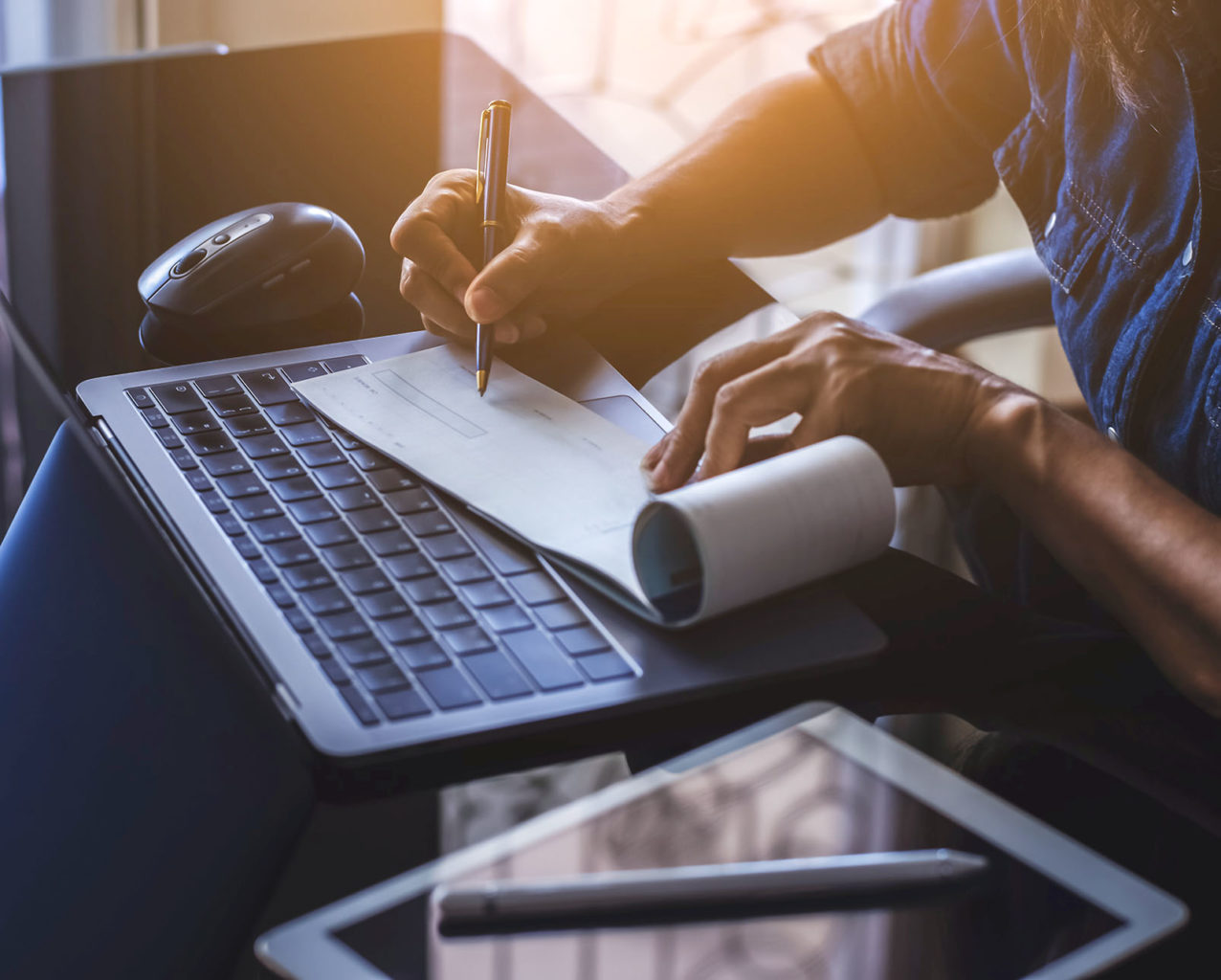 Business woman hand writing and signing white blank bank cheque book with laptop computer, mouse and digital tablet on the desk at office. Payment by check, paycheck, payroll concept.