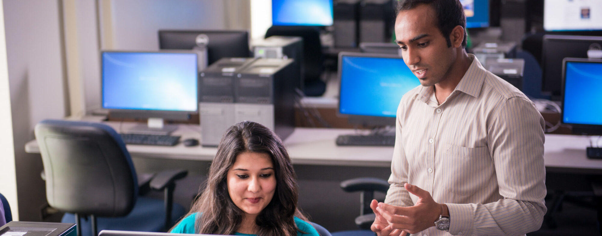 Computer science student talking with another student sitting at a computer.