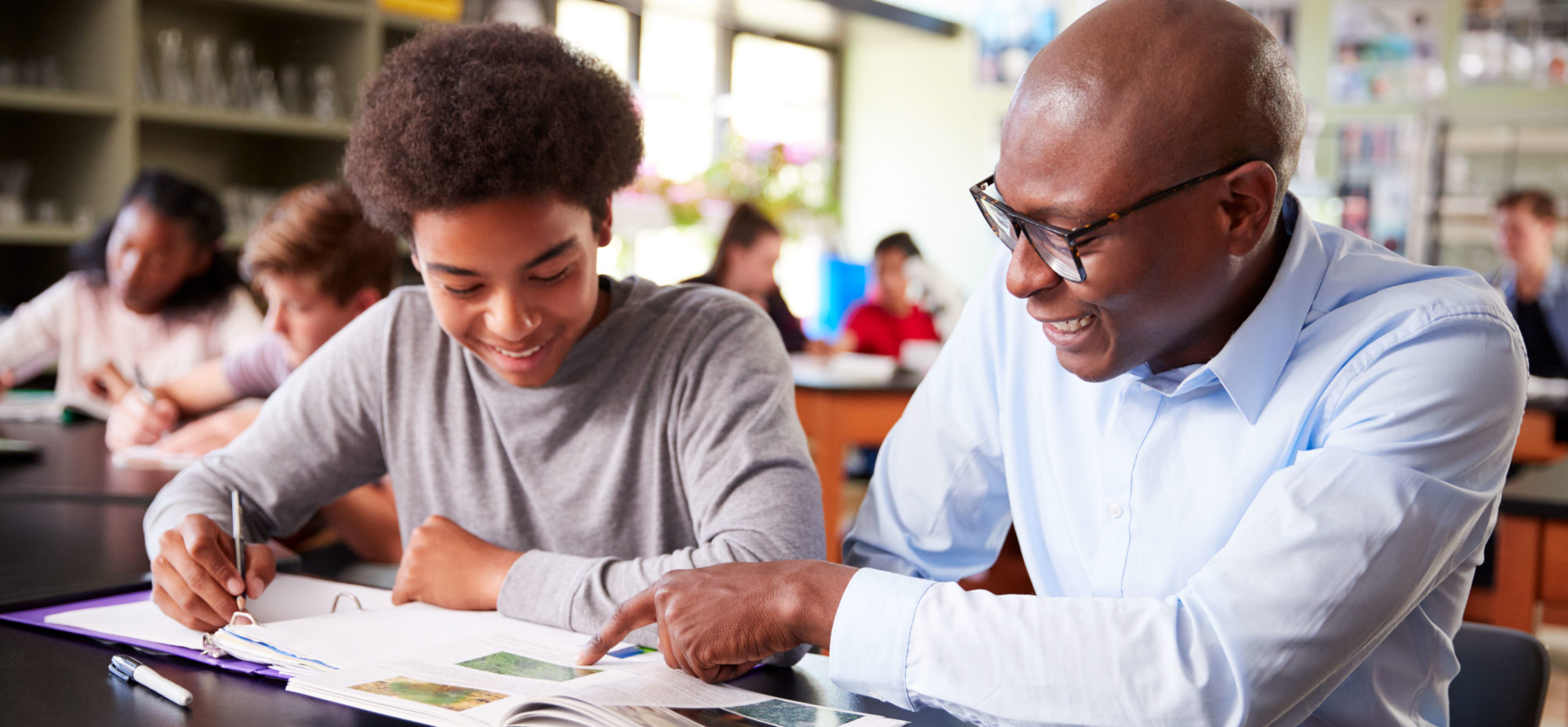 High School teacher Sitting At Desk With Male Student In Class.