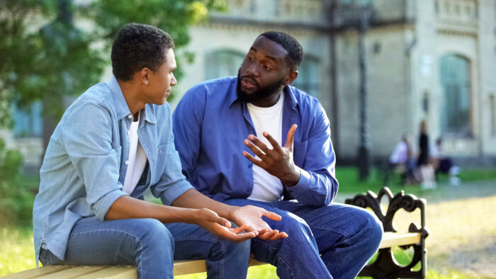 Father arguing teenage son sitting on campus bench, adolescent age difficulties.
