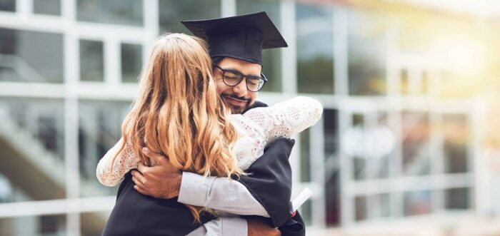 A graduate in a cap and gown happily embraces a woman with long blonde hair. They are outside, in front of a modern building with glass windows. The graduate holds a diploma in one hand, and appears joyful and proud. Sunlight shines gently in the background.