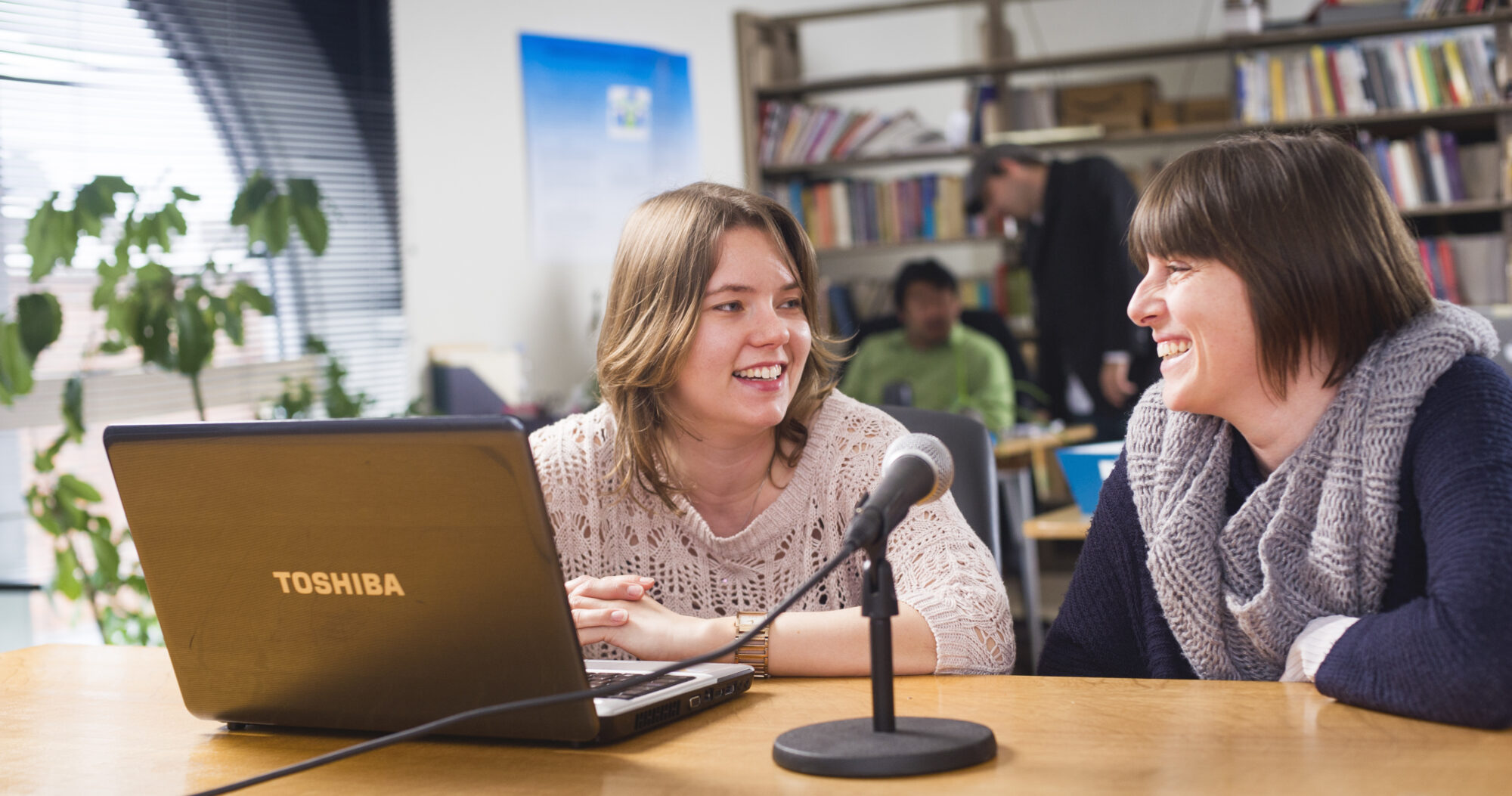 A professor and a student sitting down at a desk with a computer and a microphone in front of them.