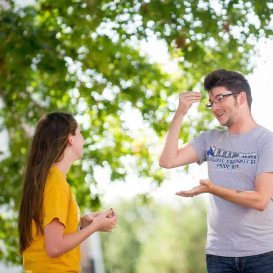 Student signing to another while under a tree.