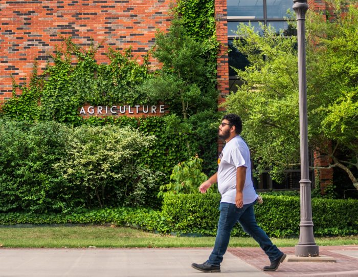 A person walking in front of a brick building with vines growing on the walls. The word "Agriculture" is spelled out in raised letters on the wall.