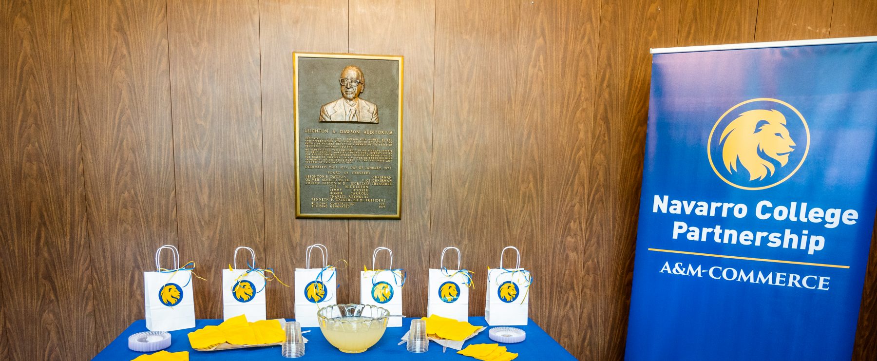 Goody bags and refreshments sit atop a table adorned with a blue and gold tablecloth next to an A&M-Commerce Navarro College Partnership flag banner.