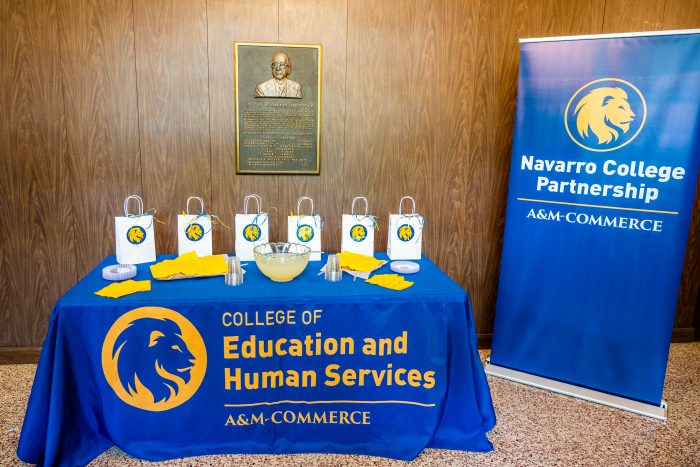 Goody bags and refreshments sit atop a table adorned with a blue and gold tablecloth next to an A&M-Commerce Navarro College Partnership flag banner.