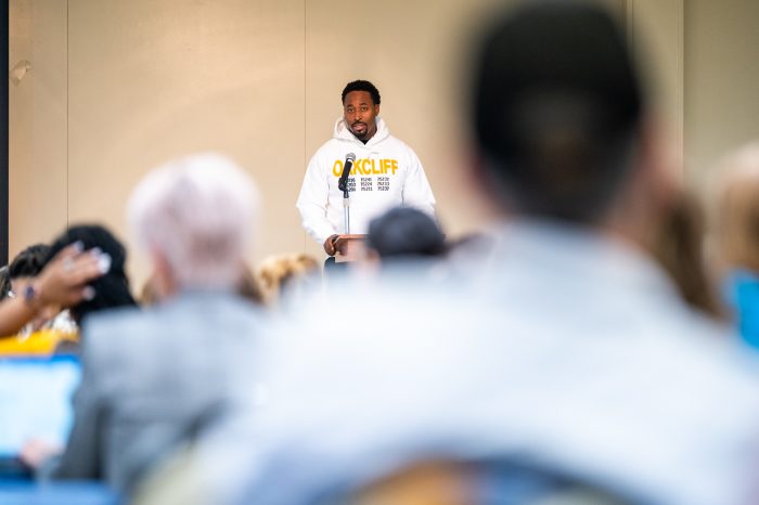A speaker wearing a white "Oak Cliff" hoodie stands at a podium, addressing an audience.