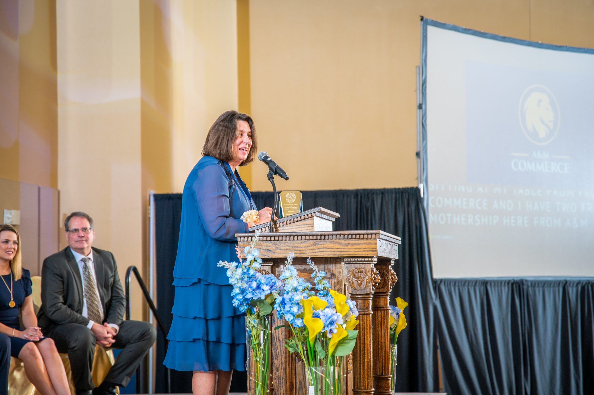 Awardee speaks to the crowd behind a podium, with two presenters sitting in chairs to her left.