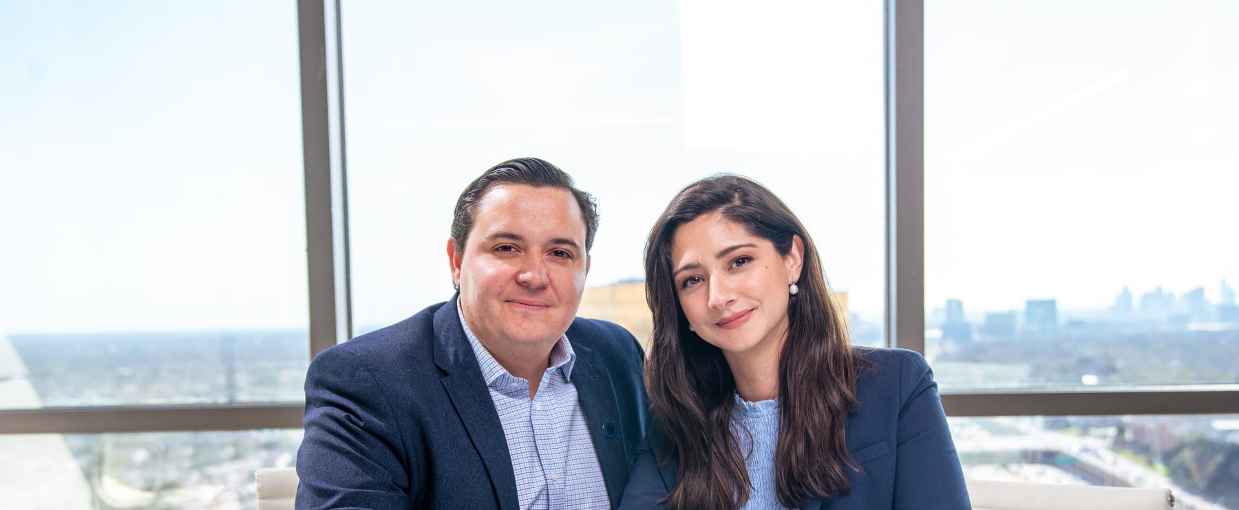 Couple poses for photo in front of glass walls with Dallas cityscape outside. Table in front of them includes their signed gift agreement.