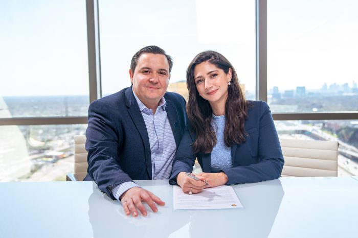 Couple poses for photo in front of glass walls with Dallas cityscape outside. Table in front of them includes their signed gift agreement.