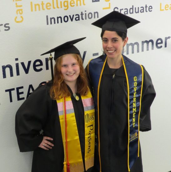 Two students, each dressed in a cap and gown, pose against a word-cloud backdrop.