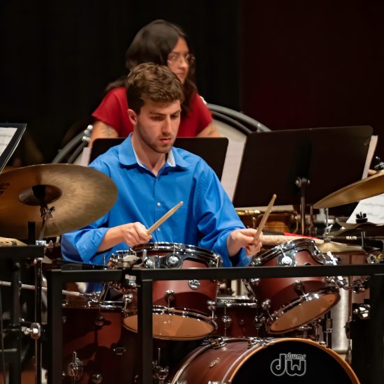 A person plays a drumset while another person plays a percussion instrument in the background.