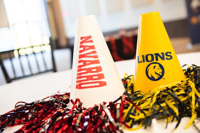 Two cheer megaphones, one white with red letters spelling NAVARRO, and one yellow with the TAMUC lion logo and the word LIONS in blue.
