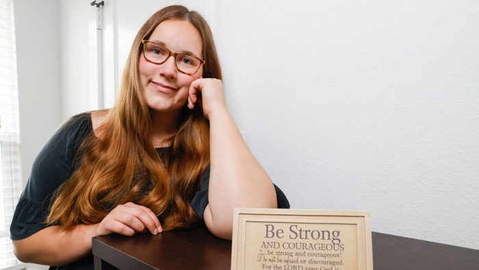 A student sits at a table, smiling with their head propped on their hand.