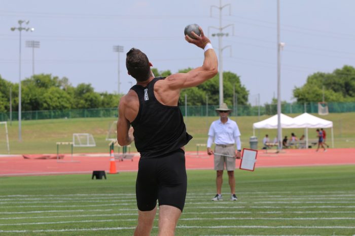 A male athlete competing in the shot put event launches a shot into the air.