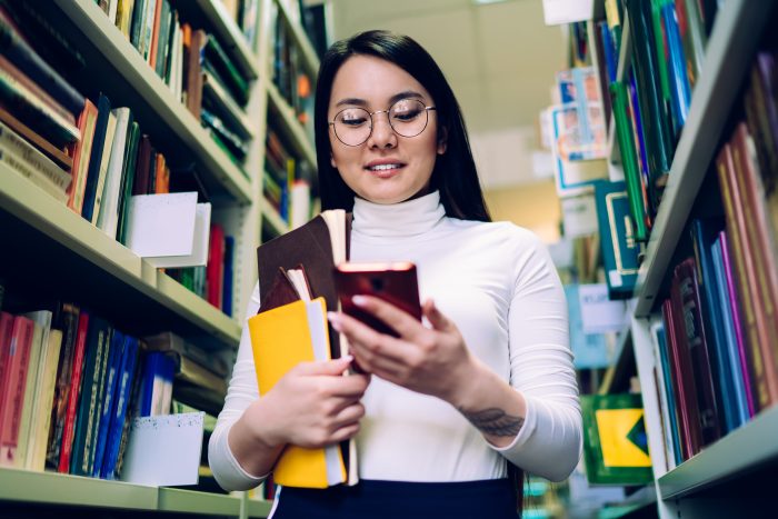 Smiling black haired young Asian female in white top and glasses with books using cellphone standing among rows of bookshelves in library
